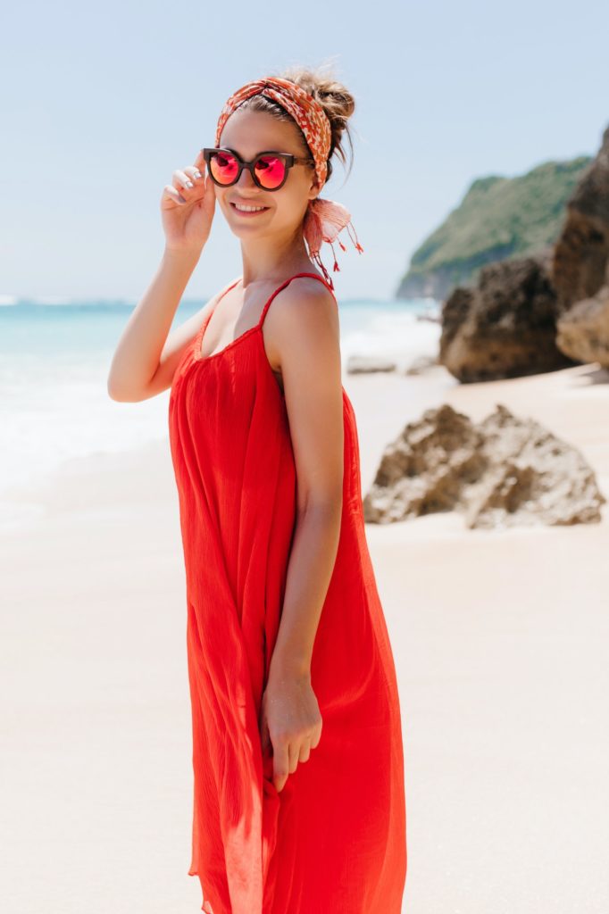 Spectacular young woman in red dress touching her sunglasses while spending time at beach. Wonderfu